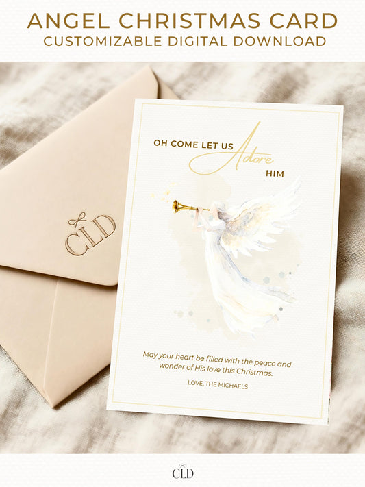 Elegant watercolor angel Christmas card with golden trumpet and text reading ‘Oh Come Let Us Adore Him,’ displayed with beige CLD envelope on soft neutral fabric background