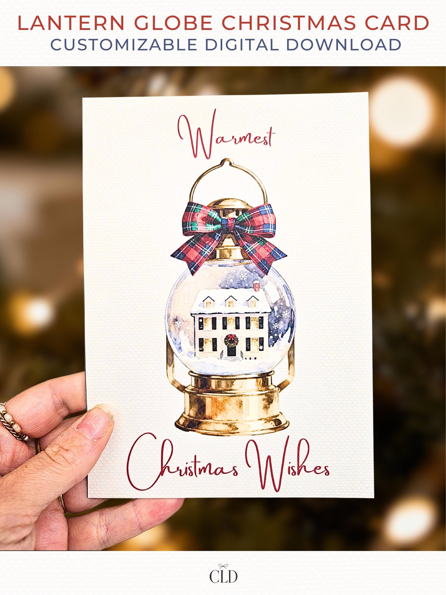 Photograph of a person holding a printed Christmas card showing a watercolor lantern snow-globe design with a red and green tartan bow, a warm neutral background with soft blurred bokeh lights, text reading "Warmest Christmas Wishes", and a subtle CLD logo at the bottom.