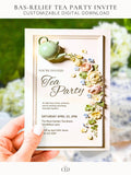 Printable tea party invitation held in hand, featuring a sculpted green teapot pouring a cascade of bas-relief flowers in blush, cream and blue. Editable 5x7 digital template for bridal shower, baby shower or brunch.